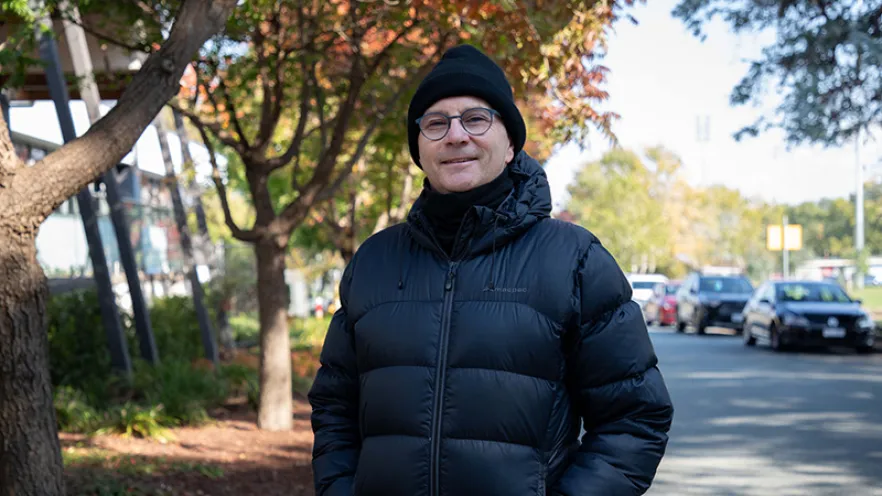 Professor Paul Kelly wearing a beanie and black puffer jacket surrounded by autumn trees standing in a street