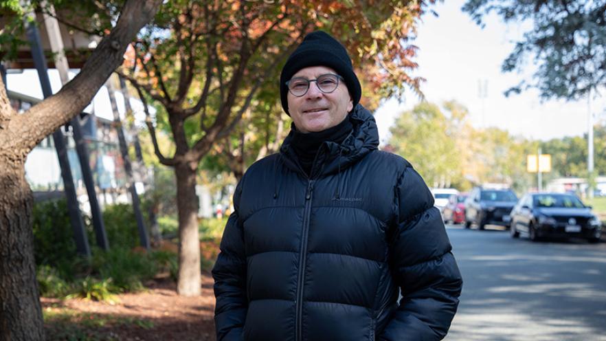 Professor Paul Kelly wearing a beanie and black puffer jacket surrounded by autumn trees standing in a street